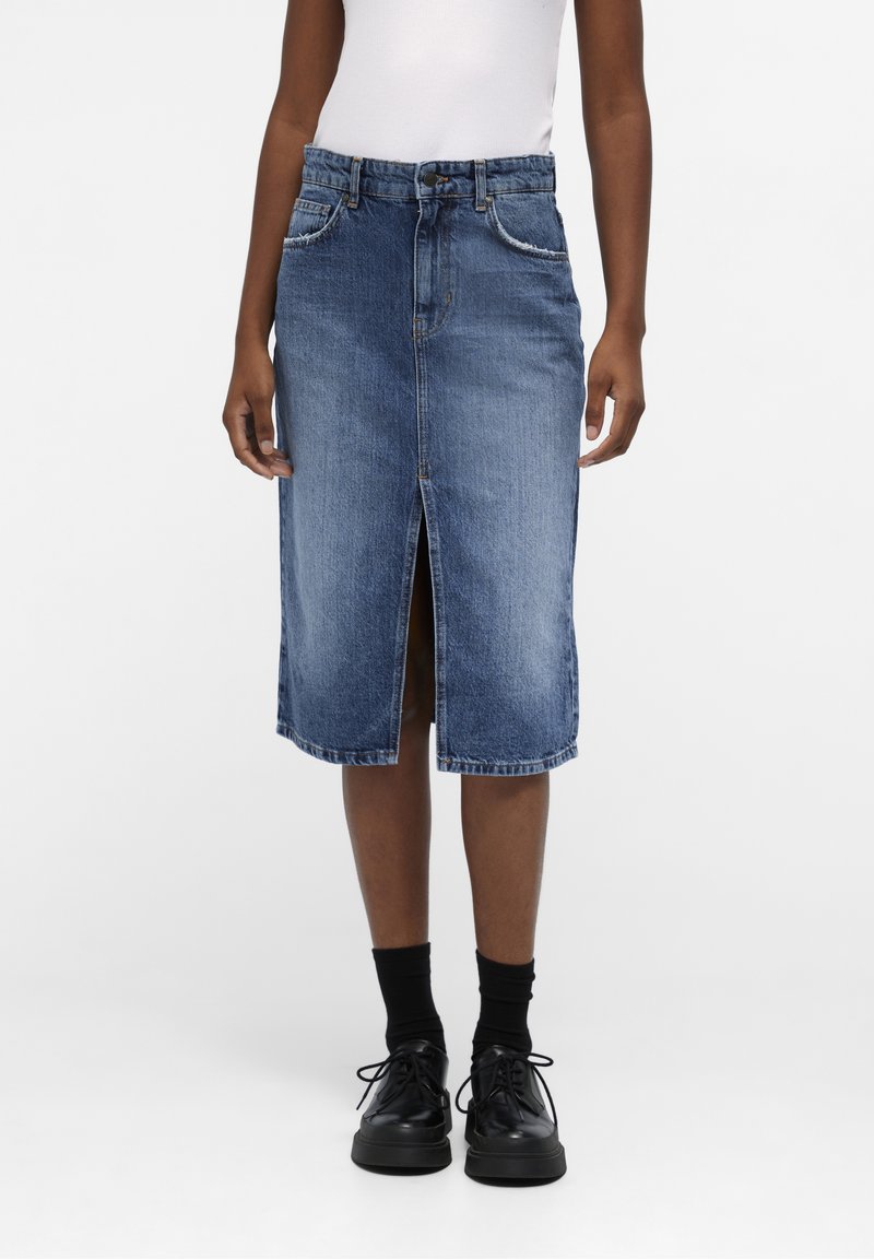 Personne portant une jupe en jean bleu longueur genou avec une fente à l'avant, un haut blanc, des chaussettes noires et des chaussures noires à lacets, debout devant un fond uni.