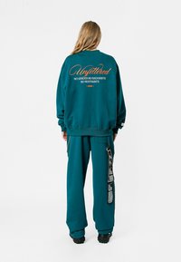 Sudadera de gran tamaño en color teal con texto bordado en la parte posterior, combinada con pantalones sueltos a juego que presentan detalles de tiras y bolsillos.