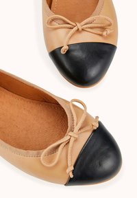 Ballerines en cuir beige avec un bout en cuir noir, présentant un détail en nœud et une texture lisse sur l'ensemble de la chaussure.