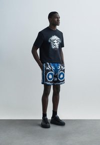 Schwarzes T-Shirt mit kurzen Ärmeln und einem grafischen Design auf der Vorderseite, kombiniert mit blauen gemusterten Shorts und schwarzen Turnschuhen.