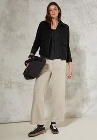 Zwarte cropped jas over een grijze cardigan, gecombineerd met crèmekleurige wijde broek en zwarte sneakers met bruine zolen. Draagt een zwarte clutch.