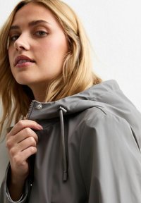 Chaqueta impermeable gris con capucha ajustable, cierre de cremallera y textura suave. El modelo sostiene la tela cerca del cuello.