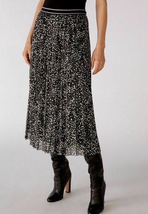 Mujer con falda midi plisada en blanco y negro con patrón abstracto y botas altas negras de tacón, de pie sobre un fondo liso.