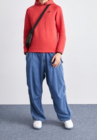 Röd sweatshirt med huva och svart logotyp, tillsammans med lösa blå jeans och vita träningsskor. Crossbody-väska bärs över axeln.