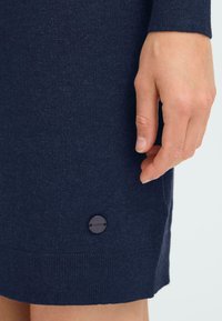 Marinblå stickad klänning med en strukturerad yta, försedd med en ribbad nederkant och en rund, svart logoknapp längst ner till vänster. Hand placerad nära nederkanten.