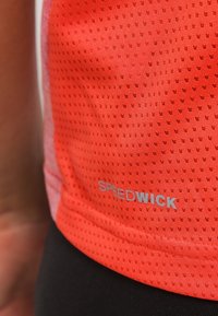 Haut de sport rouge en tissu texturé et respirant. Présente de petites perforations et un logo "SPEEDWICK" argenté sur le côté inférieur.