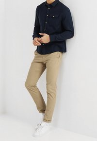 Mörkblå skjorta med knappar och ett bröstfack, kombinerad med ljusbeiga slim-fit byxor och vita sneakers på en neutral bakgrund.