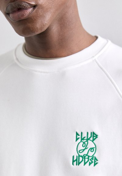 Weißer Sweater mit Rundhalsausschnitt und grünem gesticktem Textdesign, das "CLUB" über "HOUSE" in einer stilisierten Schriftart zeigt.