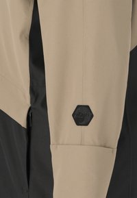 Giacca beige e nera con una texture liscia. Presenta un logo esagonale nero "SLOPE" e una tasca con zip sul lato.