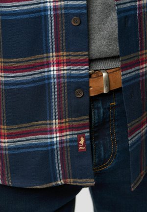 Chemise à carreaux navy avec des rayures rouges, bleues et jaunes ; boutons marron ; et une étiquette de logo. Portée par-dessus un pull gris avec un jean bleu et une ceinture marron.