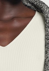 Ribbad, krämfärgad topp med V-hals, kombinerad med ett texturerat, svartvitt mönstrat yttre plagg. Mjukt material, kroppsnära passform.