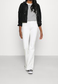 Chaqueta de terciopelo negro, blusa de manga corta a rayas, jeans blancos acampanados y zapatillas negras, con un fondo liso.