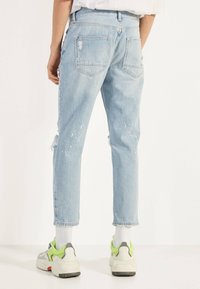 Person som bär ljusblå slitna jeans med färgfläckar, vita strumpor och vita sneakers med gröna accenter, stående mot vit bakgrund.