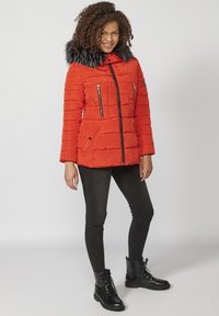 Červená puffa bunda s umelou kožušinou na kapucni, vertikálnym prešívaním, vreckami na zips a predným zipsom. Skombinovaná s čiernymi úzkymi nohavicami a topánkami.