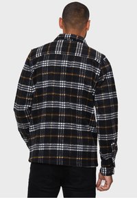Hombre que lleva una camisa de manga larga a cuadros en negro, blanco y marrón, vista desde la parte de atrás, combinada con pantalones negros.