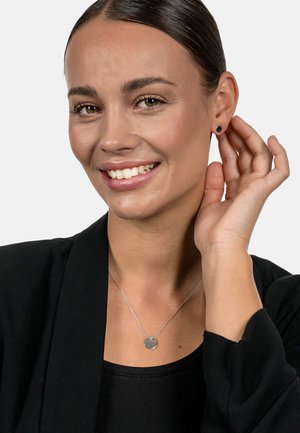 Silberne Halskette mit einem runden Anhänger, getragen über einem schwarzen Oberteil. Schwarze Steckerohrringe sichtbar; Modell lächelt und berührt ihr Ohr.