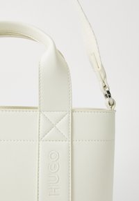 Hvit faux lær totebag med sømdetaljer, som har et merke logo og en avtakbar stropp med metalldekning.