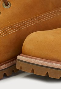 Gros plan sur des bottes en cuir beige montrant un embout arrondi, une semelle épaisse en caoutchouc avec des crampons profonds, et des coutures blanches détaillées près des lacets.