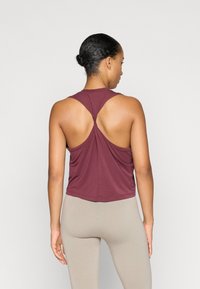 Maroon ærmeløs top med et snoet rygdesign, lavet af blødt stof. Har en afslappet pasform og en afrundet kant, parret med beige leggings.