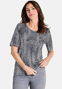 T-shirt gris à manches courtes imprimé, présentant divers motifs texturés et un col rond, fabriqué en tissu doux. Porté avec un jean gris clair.