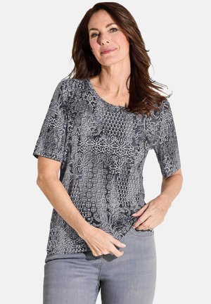 T-shirt gris à manches courtes imprimé, présentant divers motifs texturés et un col rond, fabriqué en tissu doux. Porté avec un jean gris clair.