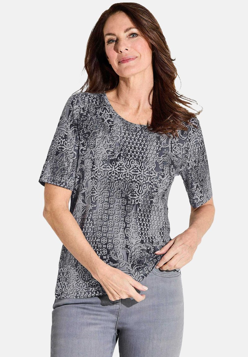 T-shirt gris à manches courtes imprimé, présentant divers motifs texturés et un col rond, fabriqué en tissu doux. Porté avec un jean gris clair.