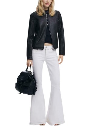 Donna con giacca di pelle nera, top corto nero, pantaloni bianchi svasati, che tiene una borsa zaino nera nella mano sinistra.