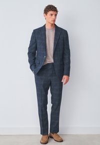 Traje a cuadros azul marino con tejido texturizado. Acompañado de un suéter de punto beige claro y zapatos color canela. Corte entallado, blazer de un solo botón.