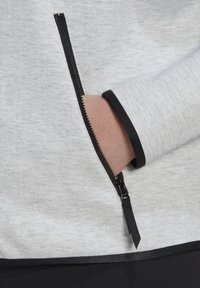 Main insérée dans la poche latérale ouverte d'un vêtement gris clair avec une fermeture éclair noire et une finition noire au niveau du poignet et de l'ourlet.