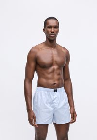 Μπλε και λευκές ριγέ σλιπ, με ελαστική μέση, κατασκευασμένες από μαλακό ύφασμα, με ετικέτα μπροστά, εμφανιζόμενες σε ανδρικό μοντέλο.