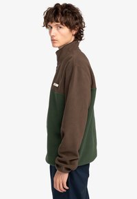 Chaqueta de forro polar marrón y verde con un cuello alto, con cierre de cremallera y una pequeña etiqueta de logotipo en la parte superior del lado izquierdo del pecho.