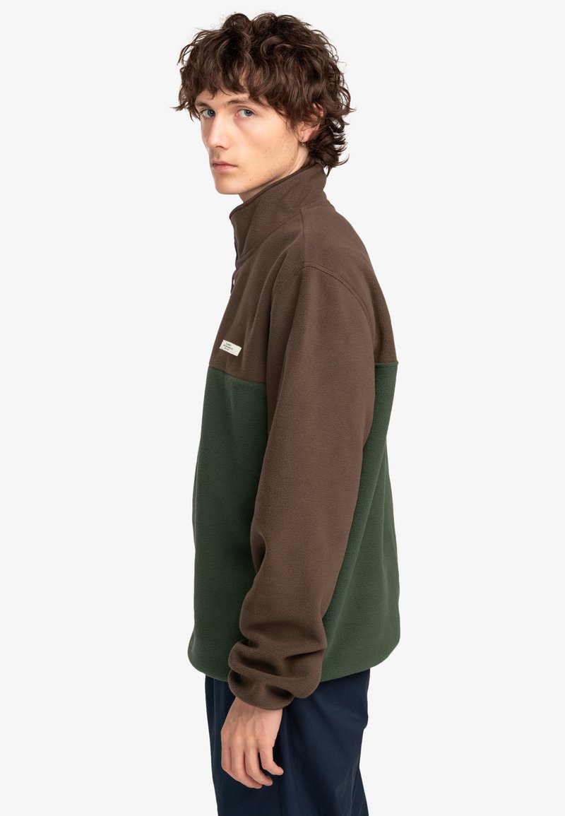 Veste en polaire marron et verte avec un col haut, dotée d'une fermeture éclair et d'une petite étiquette logo sur le haut de la poitrine gauche.