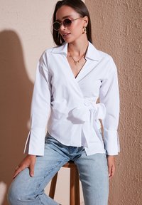 Woman wearing round sunglasses, white wrap blouse with bow, light blue jeans, gold earrings, and necklace, sitting on a wooden stool.