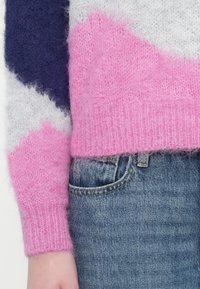 Detailní záběr na růžový, bílý a námořnický pletený svetr s chloupky, jeho rukáv a lem, doplněný o kapsu z světle modrých džínů.