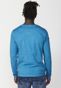 Long-sleeve shirt in vibrant blue, made from a textured fabric. Features a round neckline and subtle stitch detailing at the shoulders.