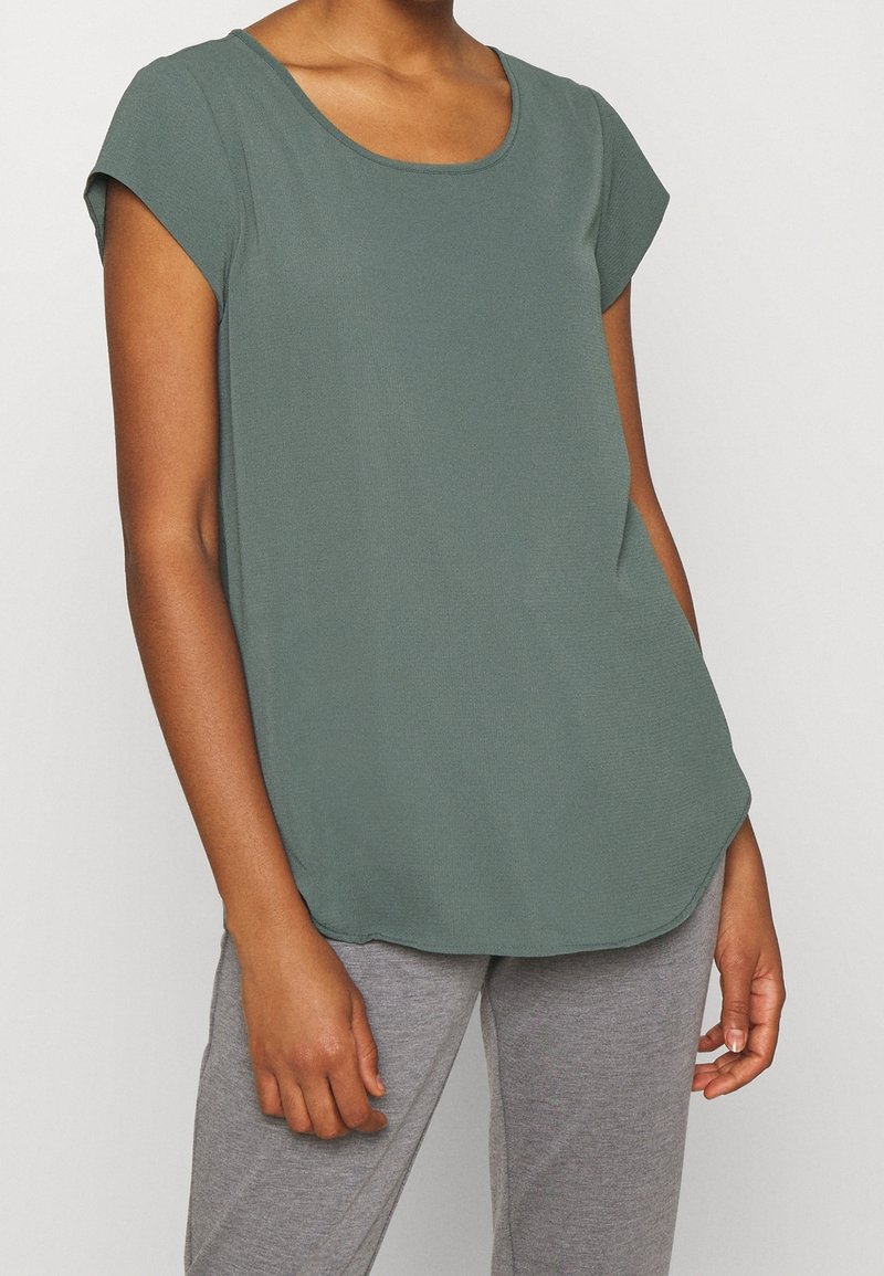 Zielona koszulka z krótkim rękawem, okrągłym dekoltem, miękką teksturą, zaokrąglonym dołem i luźnym krojem, zestawiona z szarymi spodniami.