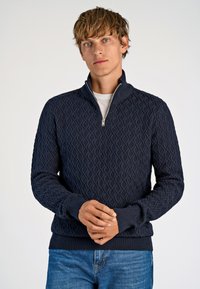 Marine gebreide trui met een ritskraag, voorzien van een gestructureerd ruitpatroon, geribbelde manchetten en zoom. Gedragen over een wit shirt.
