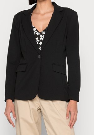 Personne portant un blazer noir à un seul bouton sur un top floral noir et blanc avec un pantalon beige, debout devant un fond uni.