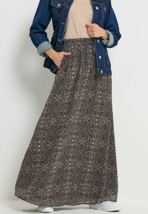 Femme portant une jupe longue à motifs, un haut beige, une veste en denim bleue aux manches retroussées et des baskets blanches, debout les mains dans les poches.