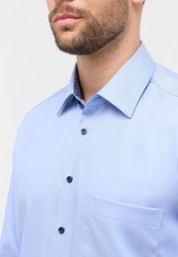 Camicia azzurro chiaro con colletto strutturato, bottoni neri e taschino sul lato sinistro. Il colletto interno presenta un motivo a contrasto.