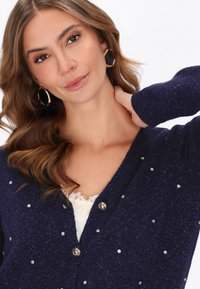 Cardigan bleumarin cu un design în formă de V, decorat cu mici strassuri. Prezintă un tricot texturat și două butoane cu ton argintiu în față.