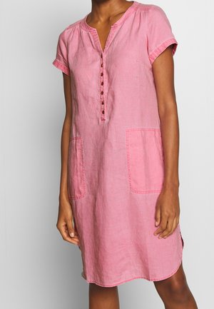 Vrouw met een roze linnen jurk met korte mouwen, knopen aan de voorkant en twee grote zakken voor, staande tegen een effen achtergrond.