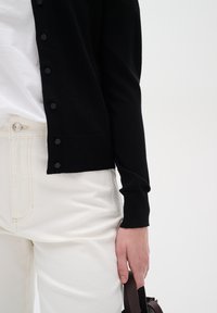 Person som bär en svart knappad kofta över en vit skjorta och vita byxor, håller en mörk väska vid sin sida mot en enfärgad bakgrund.