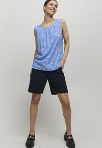Haut sans manches bleu clair avec un motif floral et des plis, associé à un short bleu marine foncé. Le mannequin porte des mocassins noirs avec des accents dorés.