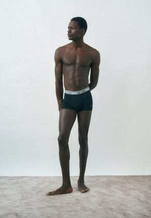 Ψηλός άνδρας, όρθιος ξυπόλητος σε δαπεδοστρωμένο πάτωμα, φορώντας μαύρα εσώρουχα Nike boxer, κοιτάζει αριστερά με τα χέρια πίσω από την πλάτη.