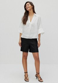 Vit linneskjorta med V-ringning och vida ärmar, i kombination med svarta shorts, som visar en avslappnad passform och subtila texturer. Svarta sandaler med remmar.