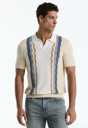 Mann trägt ein weißes Strick-Poloshirt mit kurzen Ärmeln und blauen sowie beigen vertikalen wellenförmigen Streifen, kombiniert mit ausgewaschenen blauen Jeans, steht und schaut nach rechts.