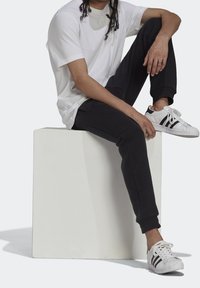 Vit bomullströja, svarta sweatpants och vita sneakers med svarta ränder. Sittande ställning med händerna vilande på knäna. Minimalistisk bakgrund.