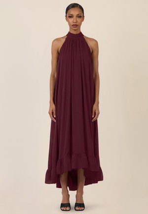 Femme debout portant une robe longue bordeaux sans manches à col montant, avec un ourlet à volants et des escarpins noirs à bout ouvert, sur un fond beige uni.