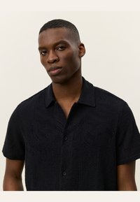 Camisa preta de manga curta feita de um tecido de malha texturizado, apresentando um padrão discreto, fecho de botões à frente e colarinho.